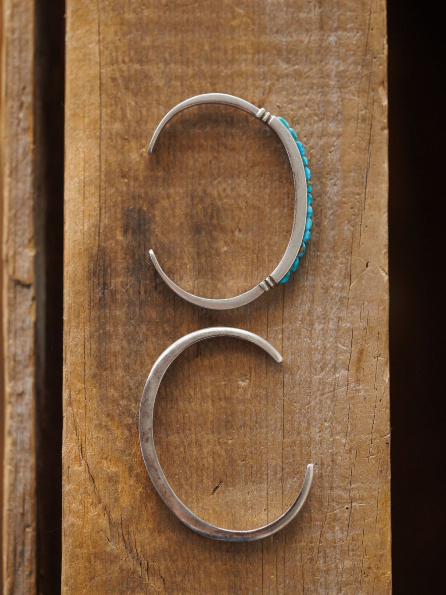 The Ponderosa Cuff by Savannah Sevens western life{&}style features two sterling silver bracelets on rustic wood: a top cuff embellished with turquoise stones and a bottom cuff showcasing a smooth, plain silver finish.
