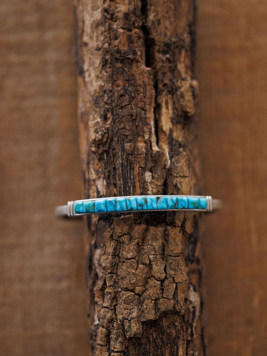 The Ponderosa Cuff by Savannah Sevens western life{&}style, a slim sterling silver bracelet with a rectangular turquoise inlay featuring natural dark veining, is displayed on rugged textured wood for striking contrast.