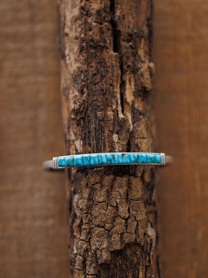 The Ponderosa Cuff by Savannah Sevens western life{&}style, a slim sterling silver bracelet with a rectangular turquoise inlay featuring natural dark veining, is displayed on rugged textured wood for striking contrast.