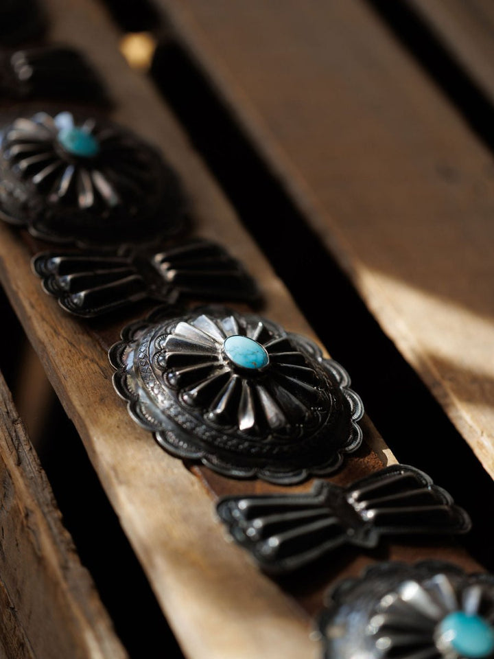 A close-up of the Portales Belt by Savannah Sevens western life{&}style, showcasing vintage sterling silver #8 Turquoise conchos with intricate engraved designs, displayed on a wooden surface in natural light.