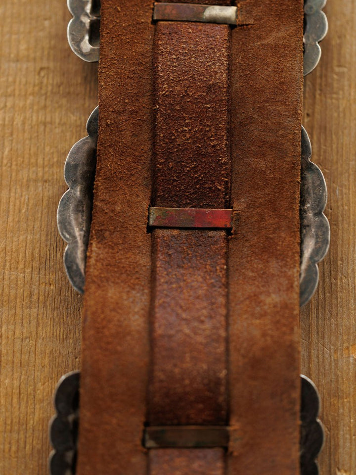 Close-up of the back side of the Portales Belt by Savannah Sevens western life{&}style, featuring worn brown leather with metal accents and scalloped edges, displayed on a wooden surface.