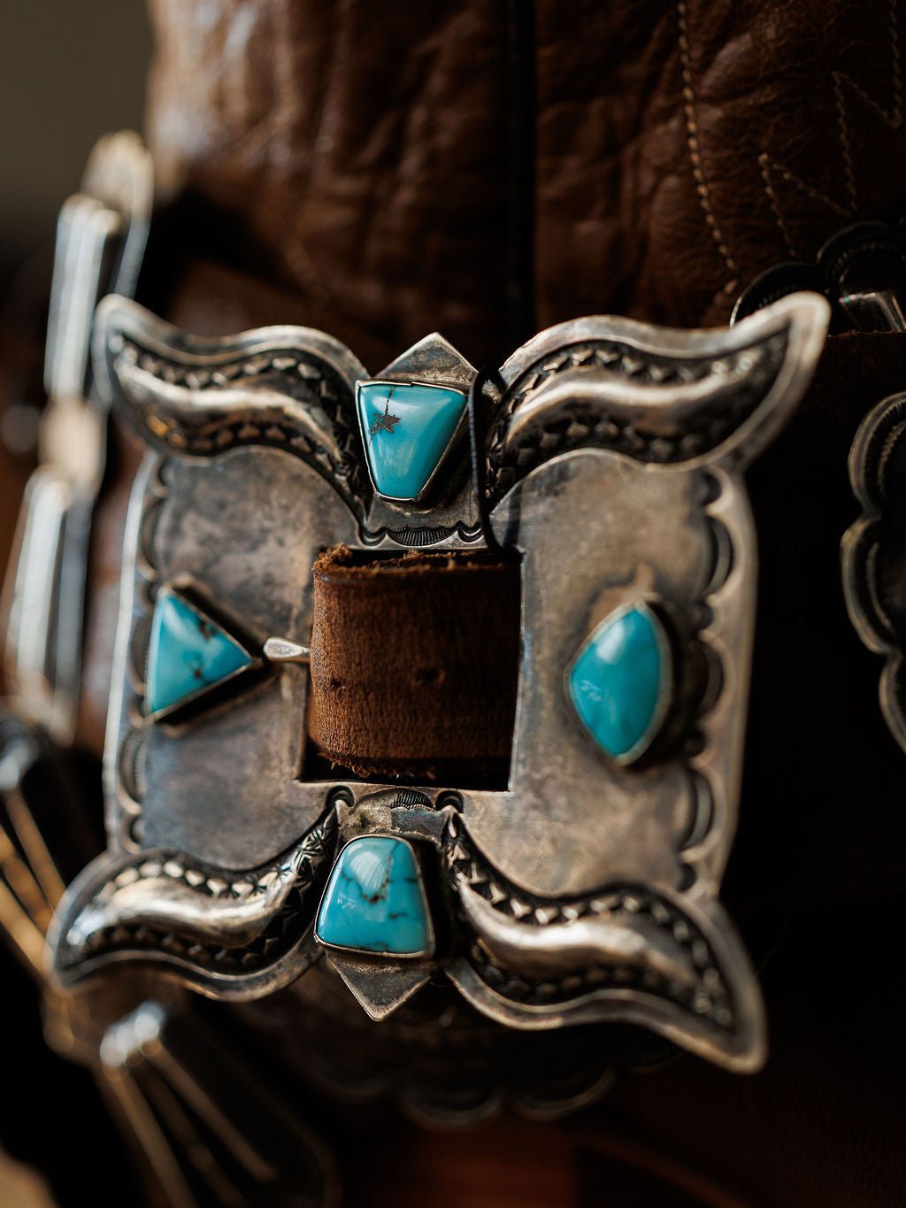 A close-up of the Portales Belt by Savannah Sevens western life{&}style, showcasing a vintage sterling silver buckle with four turquoise stones in a symmetrical design on a brown Navajo handmade leather belt.