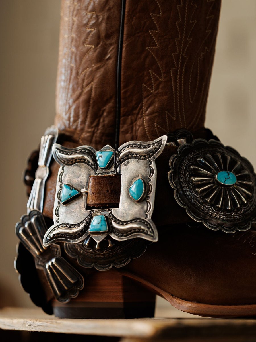 A close-up of the Portales Belt by Savannah Sevens western life{&}style, featuring brown leather with intricate Western detailing, an ornate silver buckle, and turquoise concho accents.