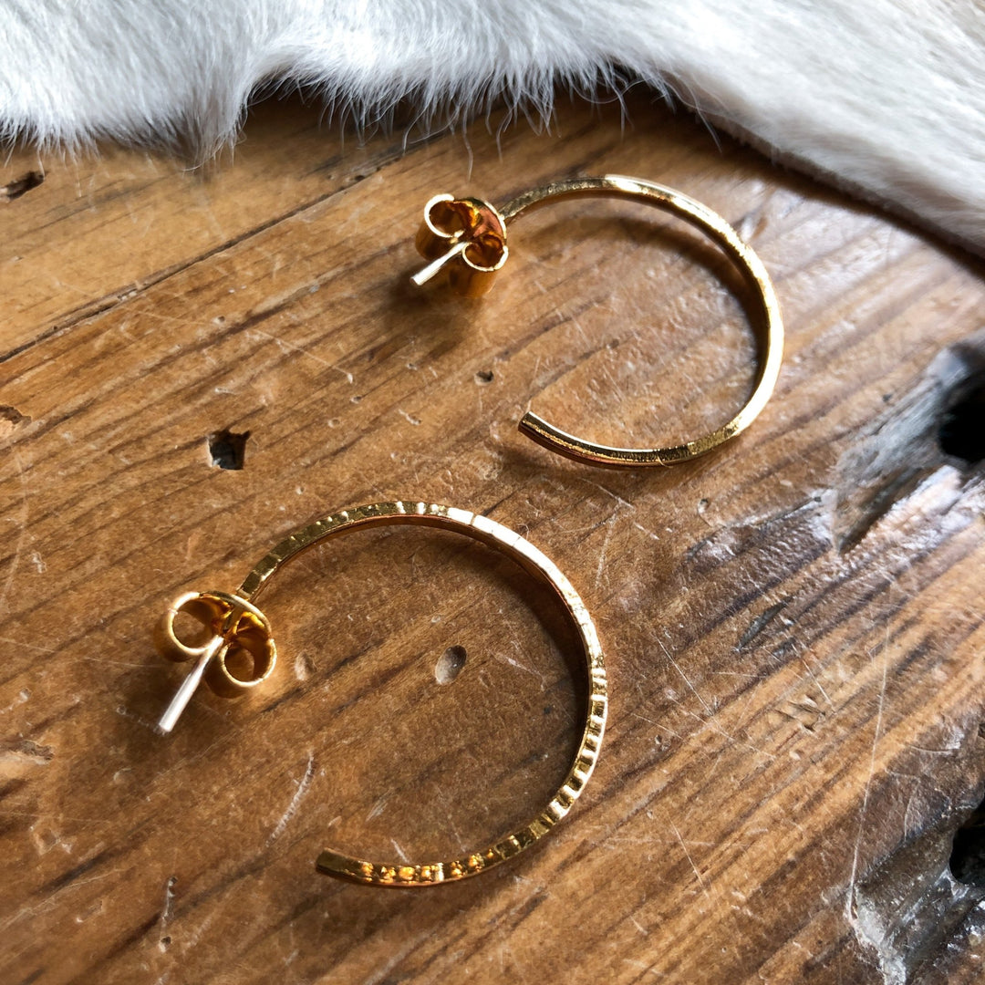 The Posen Earrings by Savannah Sevens western life{&}style—textured 24K gold-plated hoops with butterfly backs—are displayed on a rustic wooden surface beside a cowhide. 