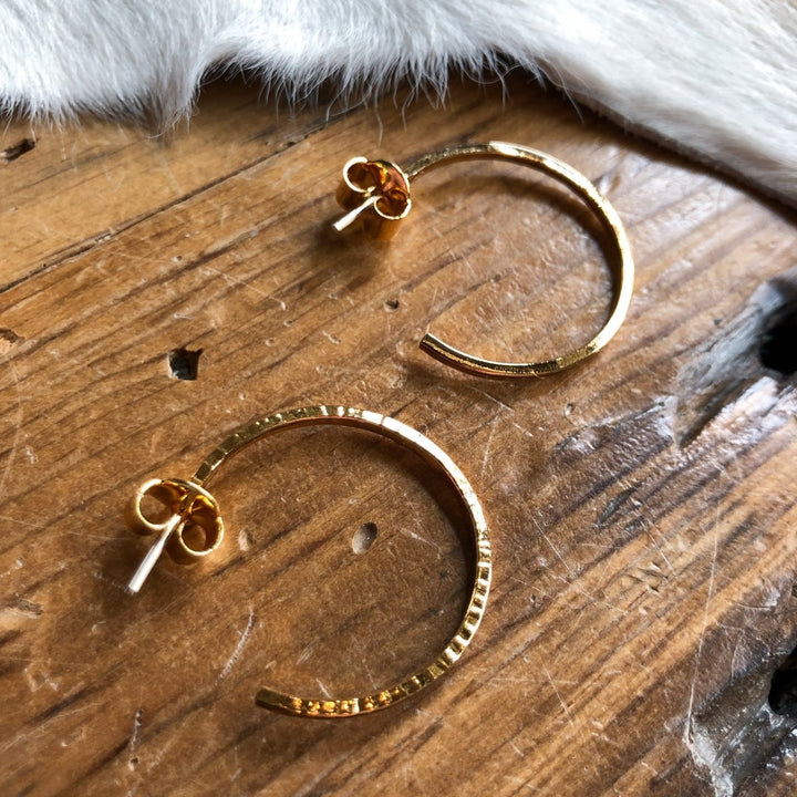 The Posen Earrings by Savannah Sevens western life{&}style—textured 24K gold-plated hoops with butterfly backs—are displayed on a rustic wooden surface beside a cowhide. 