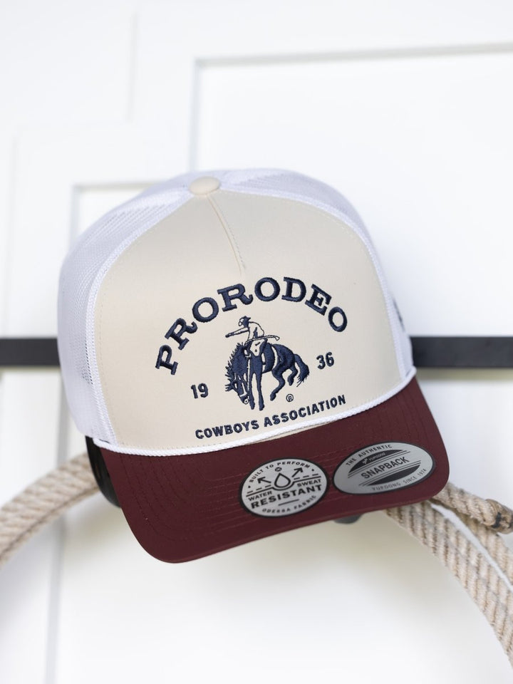 The PRCA Cap by Savannah Sevens western life{&}style is a beige and maroon trucker hat with mesh back, featuring "PRORODEO 1936 COWBOYS ASSOCIATION" above a bucking horse on a coiled rope. Water and sweat resistant.