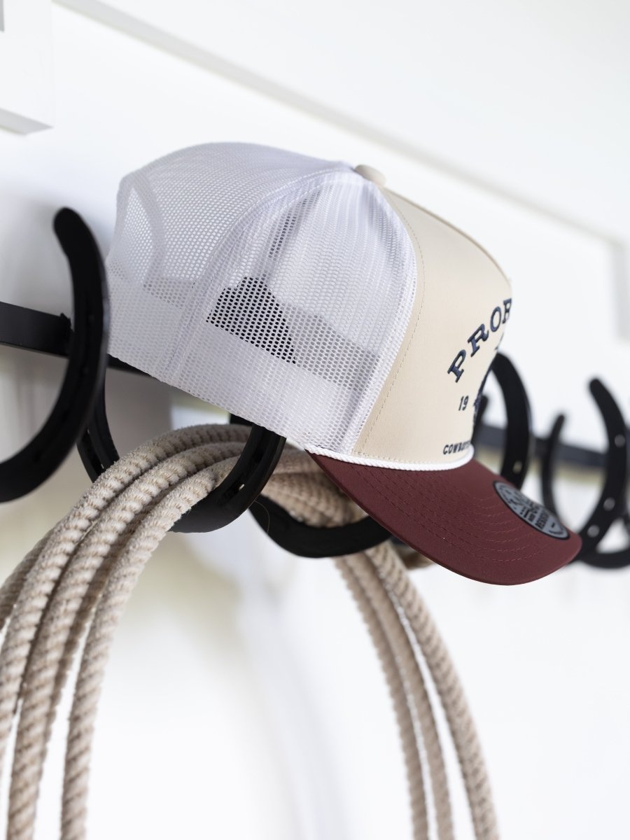 A beige and maroon Savannah Sevens western life{&}style PRCA Cap hangs on a black hook near coiled rope against a white wall. Water and sweat resistant, this cap is ideal for Pro Rodeo Cowboys Association fans.