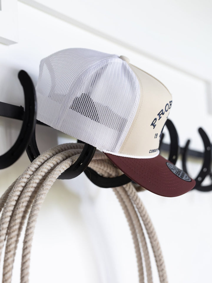 A beige and maroon Savannah Sevens western life{&}style PRCA Cap hangs on a black hook near coiled rope against a white wall. Water and sweat resistant, this cap is ideal for Pro Rodeo Cowboys Association fans.