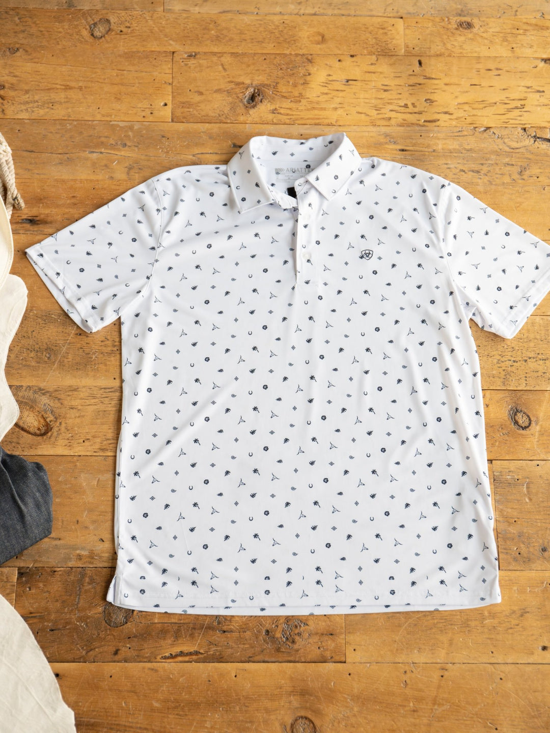 A Prescott Ariat Men's Western Graphic Polo by Savannah Sevens western life{&}style is laid flat on a wooden floor. The white short-sleeve shirt features sun protection, a collar, neckline buttons, and a small repeating navy blue pattern.