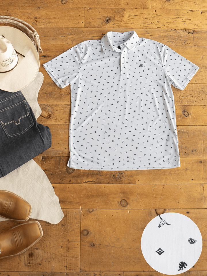 A Prescott Ariat Men's Western Graphic Polo by Savannah Sevens western life{&}style, featuring white fabric with black Western icons, is displayed flat on wood with a cowboy hat, jeans, boots, and a close-up of its moisture-wicking pattern.