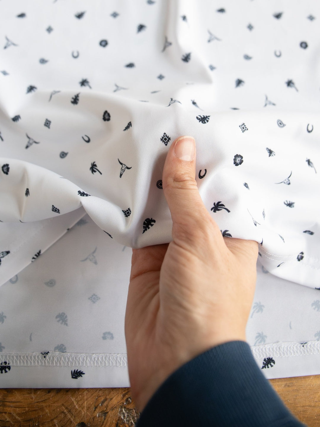 A hand holds and scrunches a white fabric with small black symbols, resembling the Prescott Ariat Men's Western Graphic Polo from Savannah Sevens western life{&}style, known for moisture-wicking, against a wooden surface.