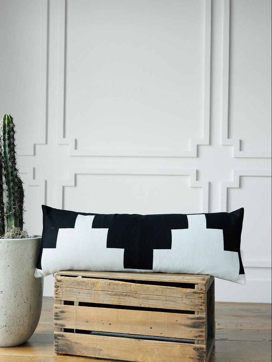 The Presidio Lumbar Pillow Cover {B & W} by Savannah Sevens western life{&}style features a luxury black and white geometric design, displayed on a wooden crate next to a cactus in a concrete pot by a white paneled wall.