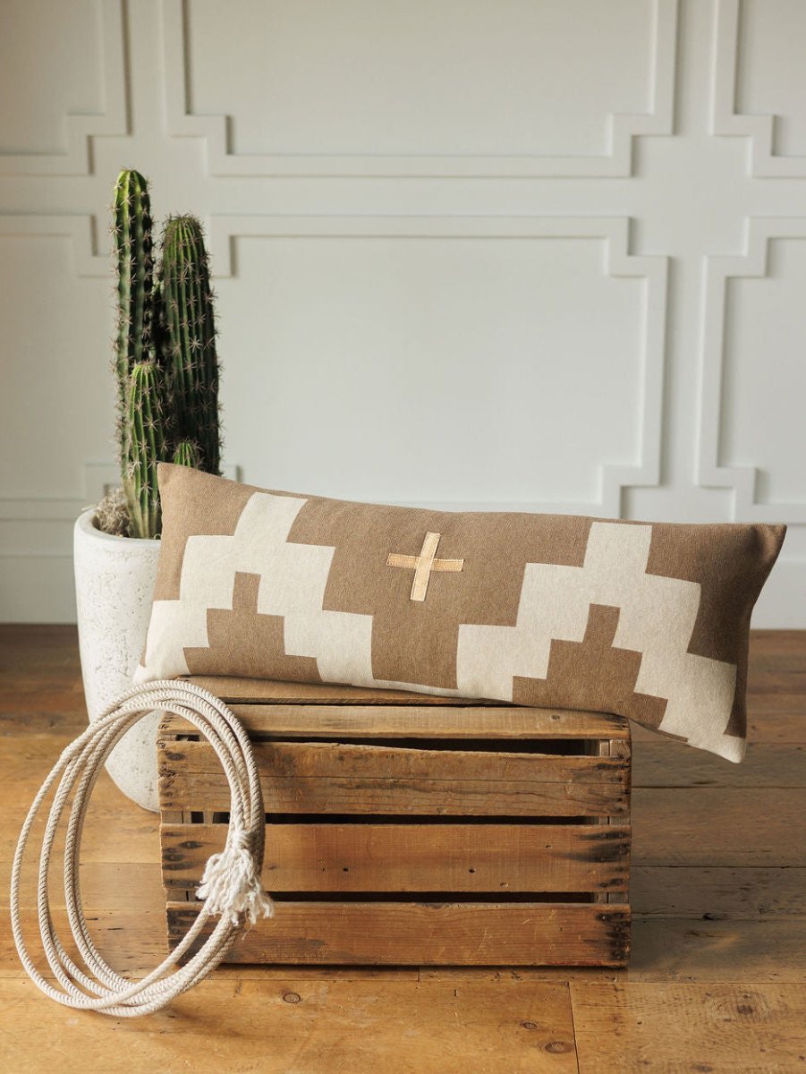 The Presidio Lumbar Pillow Cover {Tan} by Savannah Sevens western life{&}style, with a geometric cross pattern, rests on a wooden crate beside coiled white rope and a cactus in a white planter, set against a paneled wall.