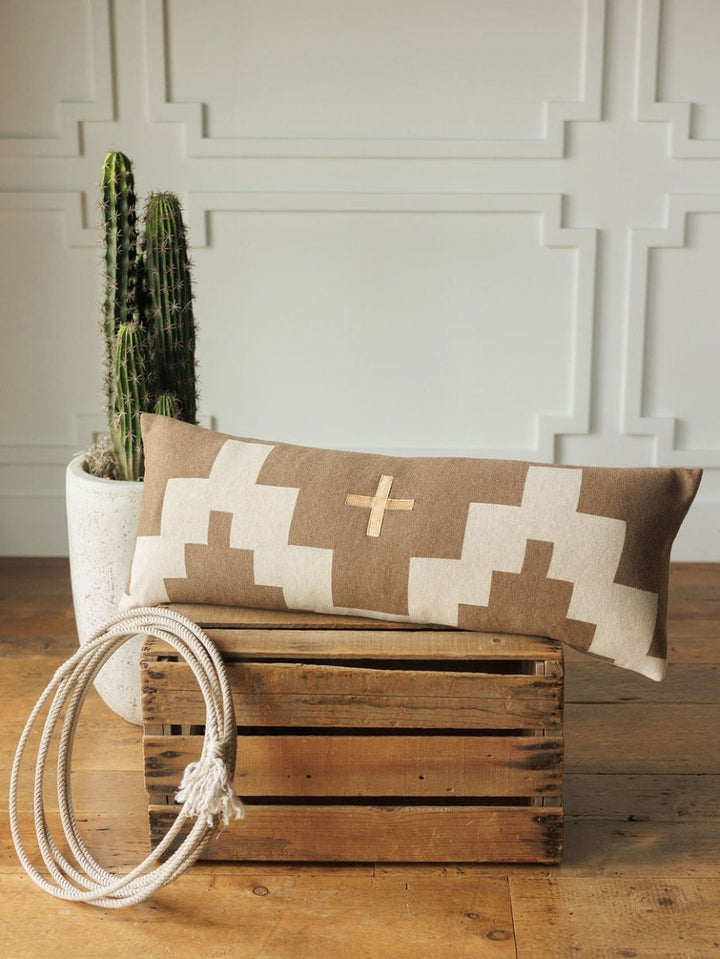 The Presidio Lumbar Pillow Cover {Tan} by Savannah Sevens western life{&}style, with a geometric cross pattern, rests on a wooden crate beside coiled white rope and a cactus in a white planter, set against a paneled wall.