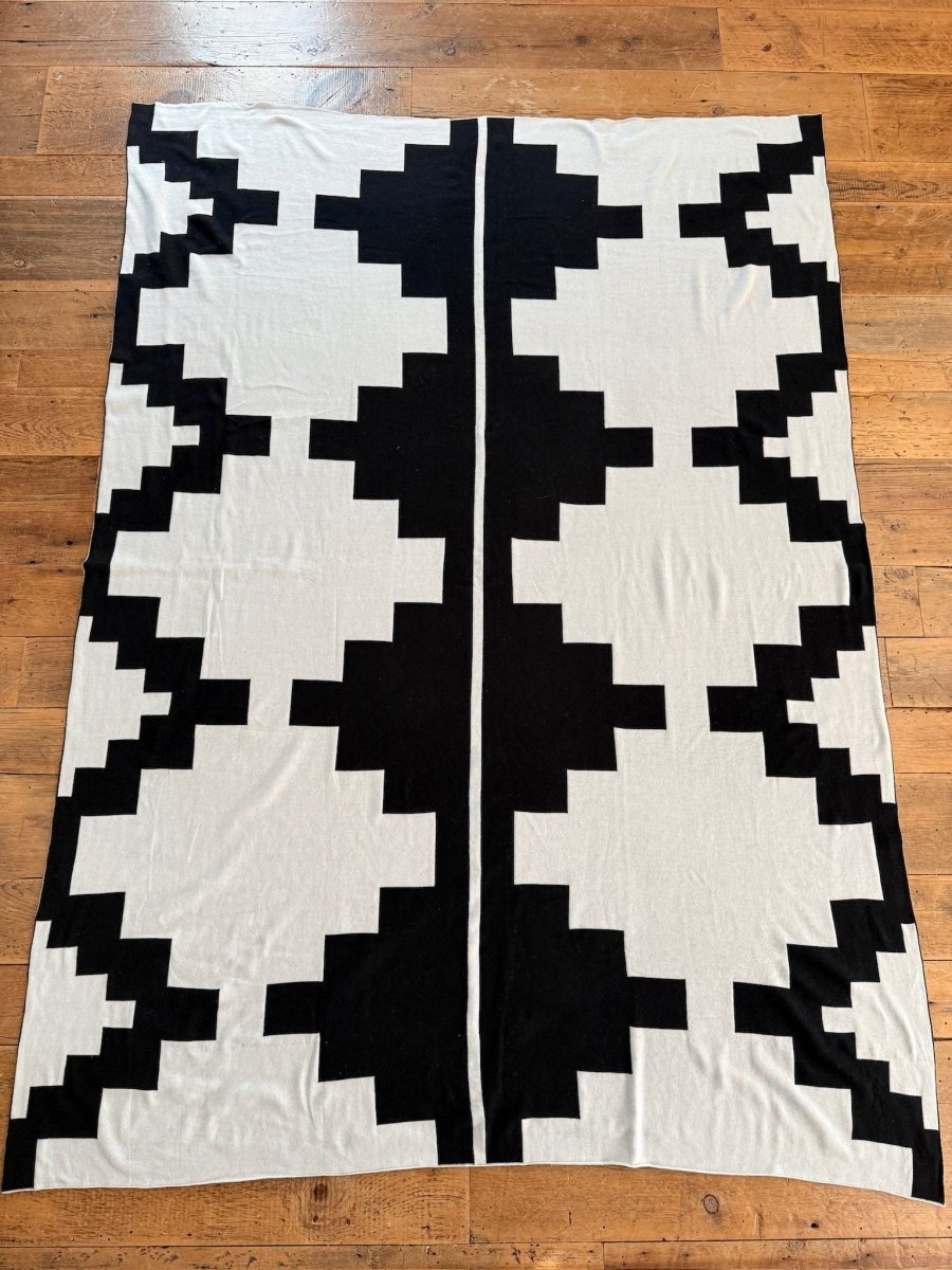 The Presidio Throw Blanket {B & W} by Savannah Sevens western life{&}style features a bold black geometric pattern on light gray, with large stepped shapes symmetrically arranged along a central line, displayed flat on a wooden floor.