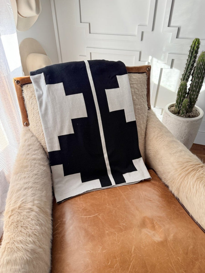 A cozy chair with a brown leather seat is draped with the Presidio Throw Blanket {B & W} by Savannah Sevens western life{&}style. A potted cactus and hats add Western Grande flair near a white paneled wall.