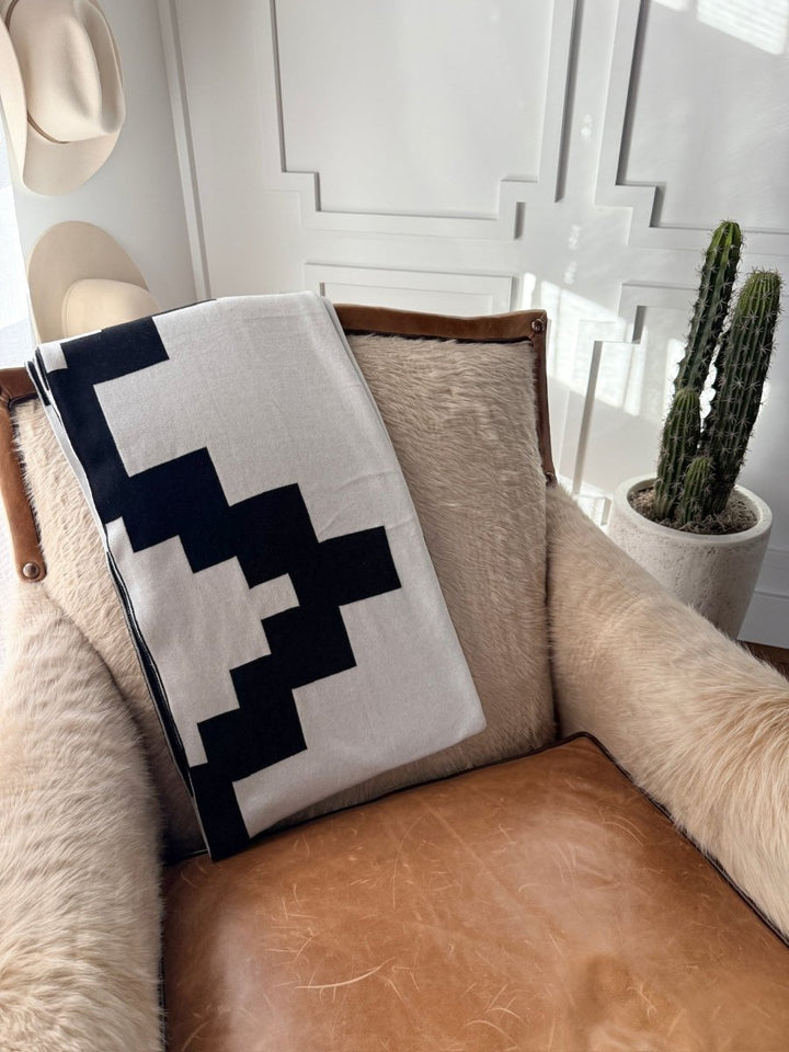 A cozy tan leather chair with Savannah Sevens western life{&}style’s Presidio Throw Blanket {B & W}, a light fur throw, a tall potted cactus, and two hats on a white-paneled wall. Sunlight adds Western Grande flair.