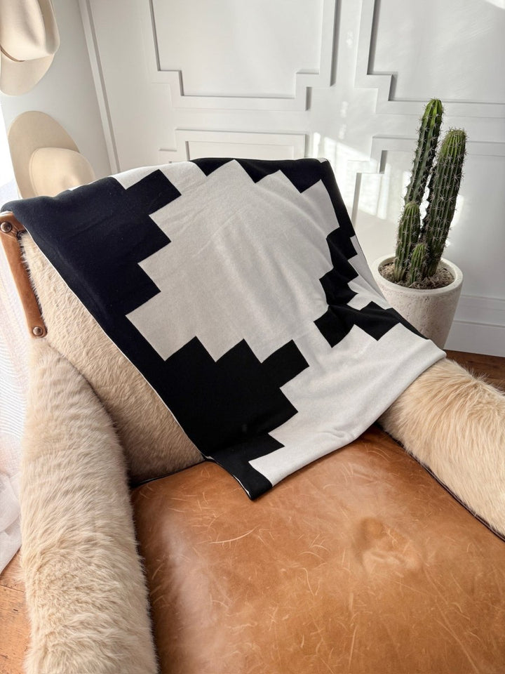 A tan leather armchair with beige cushions is draped with the Savannah Sevens western life{&}style Presidio Throw Blanket {B & W}, featuring black and white geometrics. A concrete-potted cactus nearby completes the refined Western Grande look.