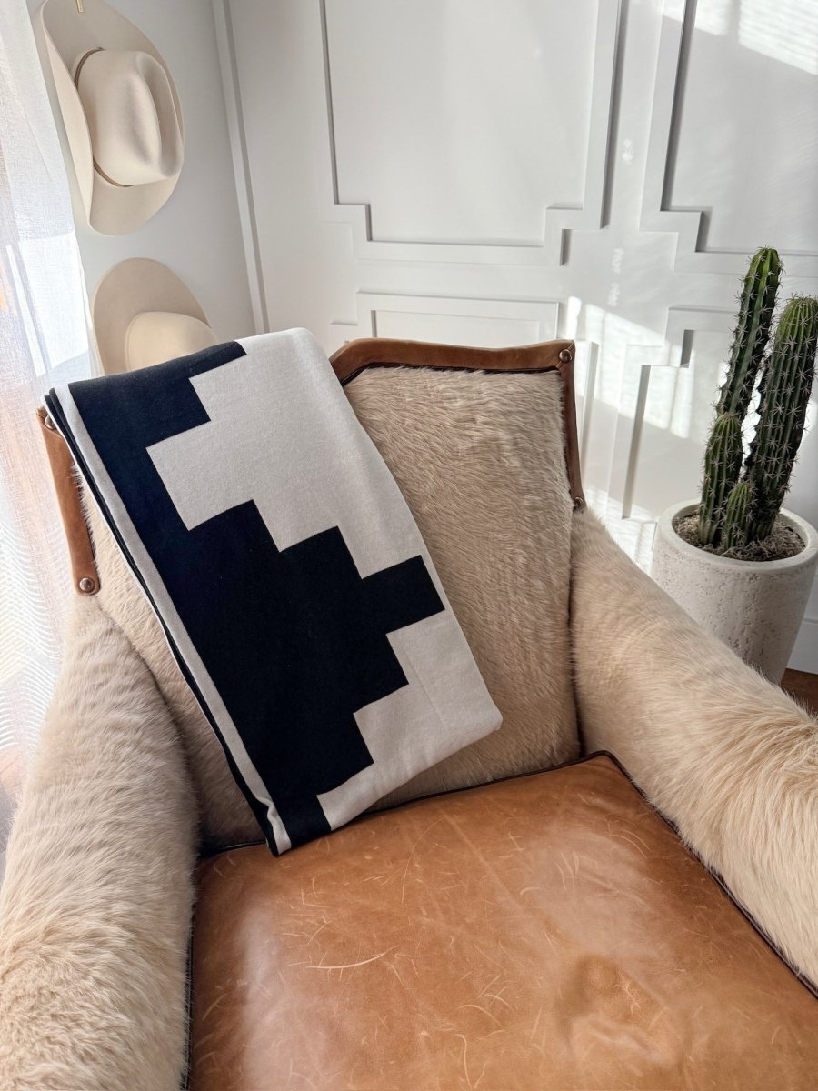 A cozy leather and fur chair features the Presidio Throw Blanket {B & W} by Savannah Sevens western life{&}style, draped over the back, beside a tall cactus in a white pot and hats on the wall in a sunlit room.