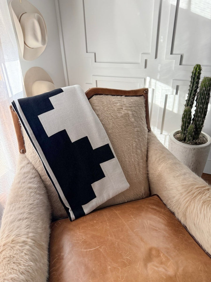 A cozy leather and fur chair features the Presidio Throw Blanket {B & W} by Savannah Sevens western life{&}style, draped over the back, beside a tall cactus in a white pot and hats on the wall in a sunlit room.