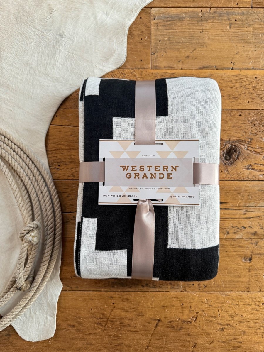 The Presidio Throw Blanket {B & W} by Savannah Sevens western life{&}style features a folded black and white geometric Southwest design, tied with satin ribbon and Western Grande label, sitting next to a white cowhide rug. Enjoy its luxury soft micro knit texture.