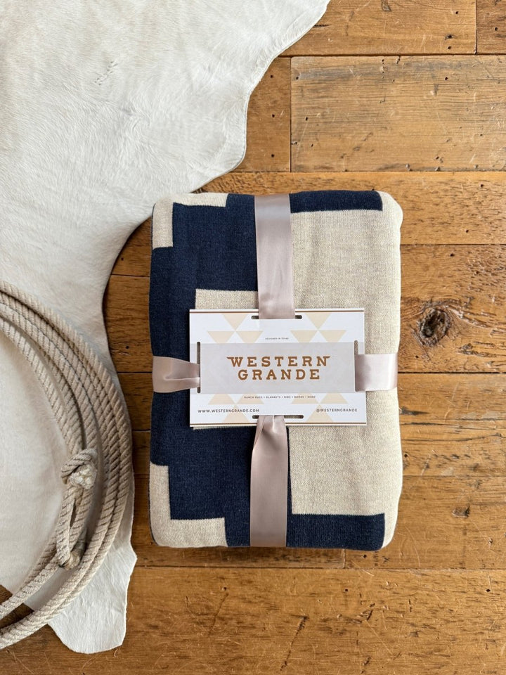 The Presidio Throw Blanket {Midnight Navy} by Savannah Sevens western life{&}style, featuring a geometric pattern, is folded with a satin ribbon and brand tag, resting on a wooden floor beside coiled rope and a white animal hide rug.