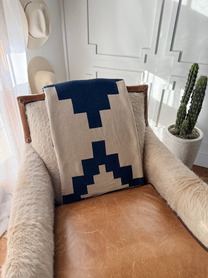 A leather and fur armchair with a Savannah Sevens western life{&}style Presidio Throw Blanket in Midnight Navy draped over the backrest sits in a sunlit room with white paneling, near a potted cactus and two hats hanging on the wall.