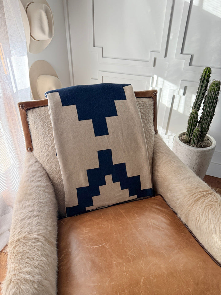 A leather and fur armchair with a Savannah Sevens western life{&}style Presidio Throw Blanket in Midnight Navy draped over the backrest sits in a sunlit room with white paneling, near a potted cactus and two hats hanging on the wall.