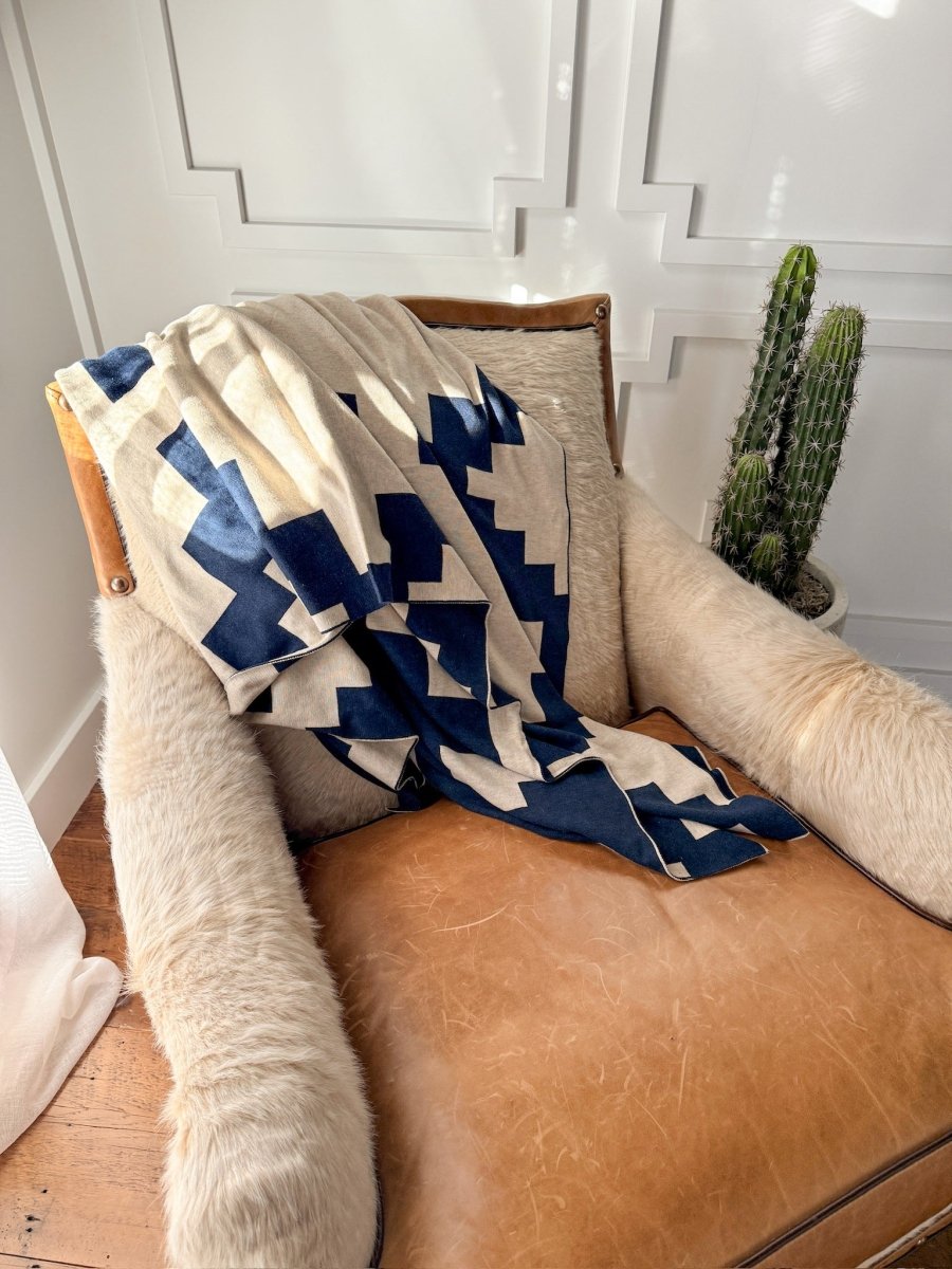 The Presidio Throw Blanket in Midnight Navy by Savannah Sevens western life{&}style is draped over a tan leather armchair beside a green cactus in a white pot, all set against a white paneled wall in a sunlit room.