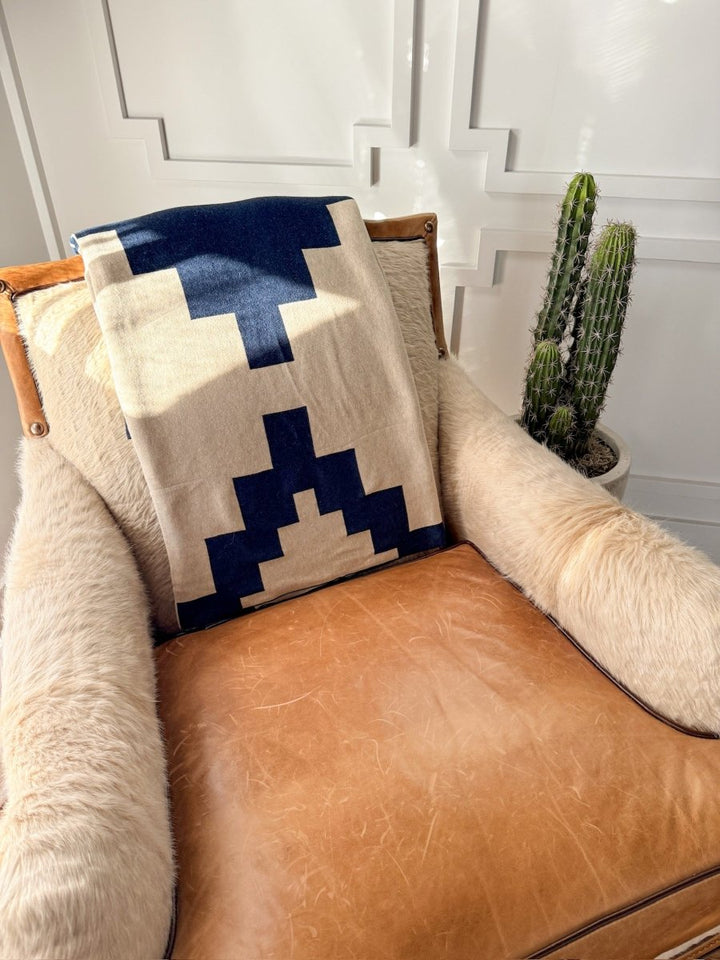 A tan leather armchair is draped with a luxury soft micro knit faux fur throw and the Presidio Throw Blanket {Midnight Navy} by Savannah Sevens western life{&}style, while a tall potted cactus sits beside it against a sunlit white paneled wall.