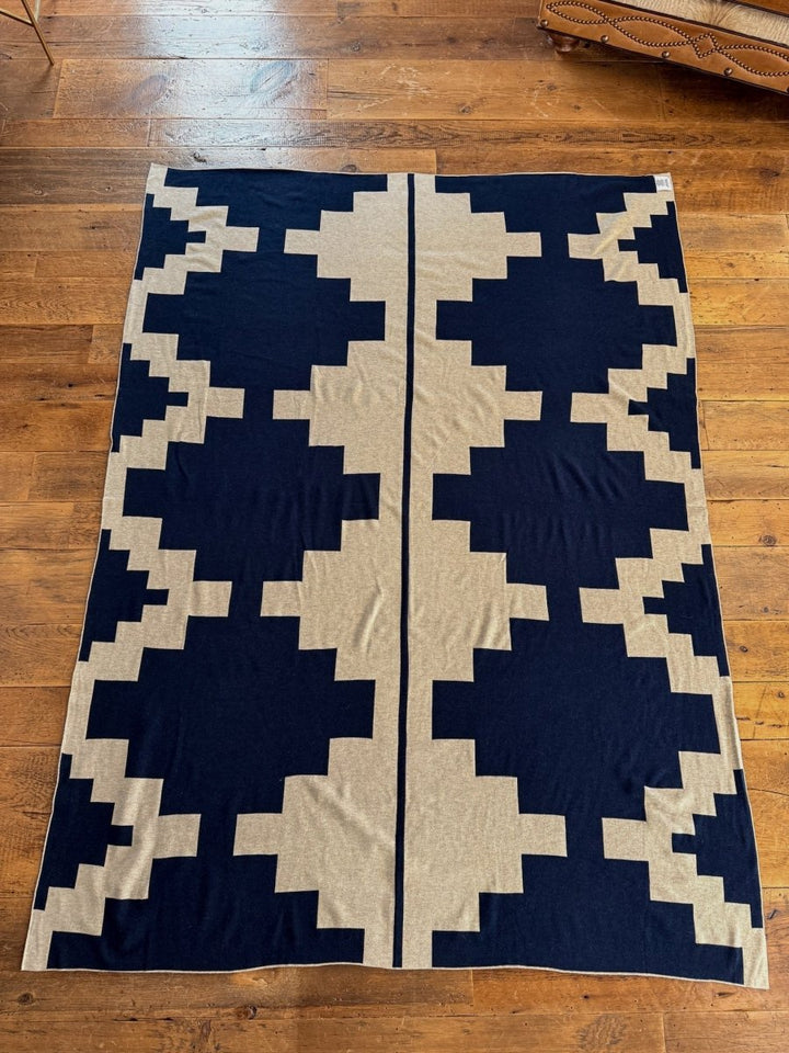 The Presidio Throw Blanket in Midnight Navy by Savannah Sevens western life{&}style features a soft, reversible micro knit with beige and dark blue geometric shapes, laid flat on a wooden floor with a symmetrical central vertical design.