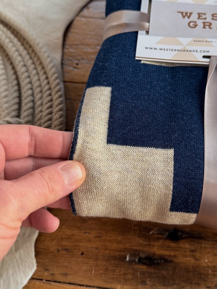A hand holds the Presidio Throw Blanket {Midnight Navy} by Savannah Sevens western life{&}style—100% cotton with a navy and beige geometric pattern, folded with a satin ribbon, on wood with cowhide in the background.
