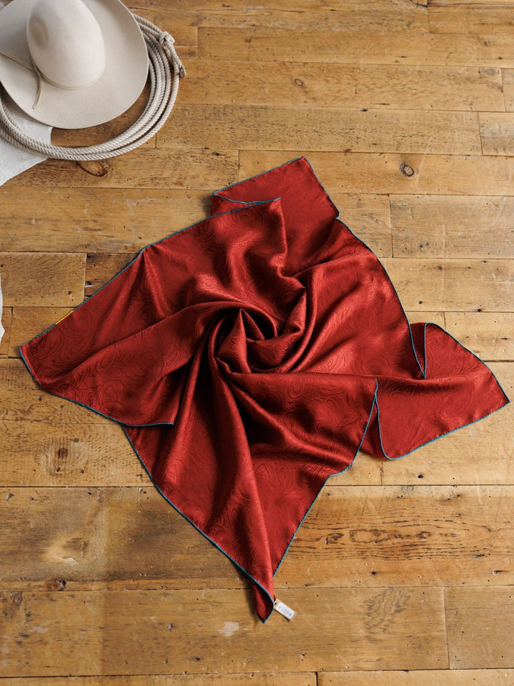 The Prime Red Paisley Silk Wild Rag by Savannah Sevens western life{&}style is styled in a spiral on a wooden floor, with a white cowboy hat, rope, and part of a white garment adding a classic western vibe in the corner.