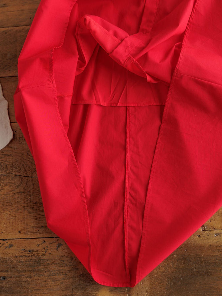 The Primera Red Cotton Dress by Savannah Sevens western life{&}style, in a vivid tomato hue, is partially folded on a wooden surface with visible stitching and seams. A light beige fabric piece is seen at the left edge of the image.