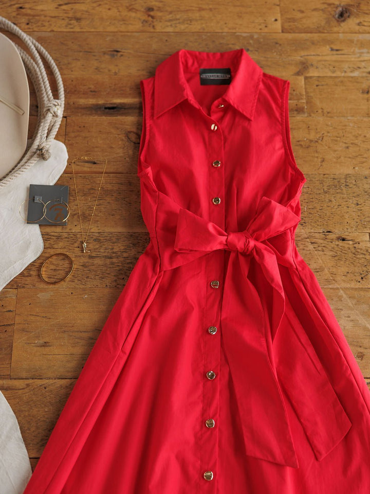 The Primera Red Cotton Dress by Savannah Sevens western life{&}style, a sleeveless tomato-red midi with a waist bow, rests on a wooden surface with gold hoop earrings, a necklace, and a beige rope-handle bag nearby.