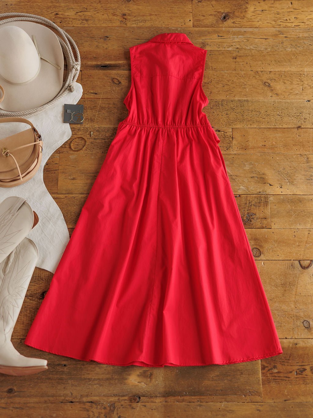 The Primera Red Cotton Dress by Savannah Sevens western life{&}style, featuring a cinched waist and adjustable back tie, is laid flat on a wooden floor with white cowboy boots, a tan purse, a white hat, and a white garment.