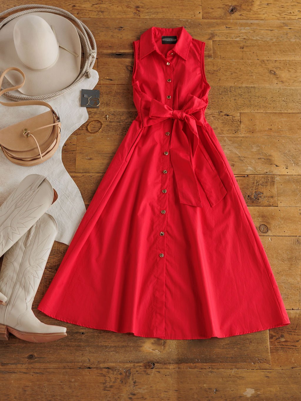 The Primera Red Cotton Dress by Savannah Sevens western life{&}style is laid flat on a wooden floor, styled with white cowboy boots, a beige hat, a small tan purse, and a white folded sweater.