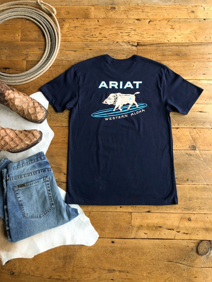 A navy Pua'a Ariat Men's Graphic Tee by Savannah Sevens western life{&}style, featuring a white bull and “Western Aloha” text, lies flat on a wooden floor beside brown cowboy boots and blue jeans.