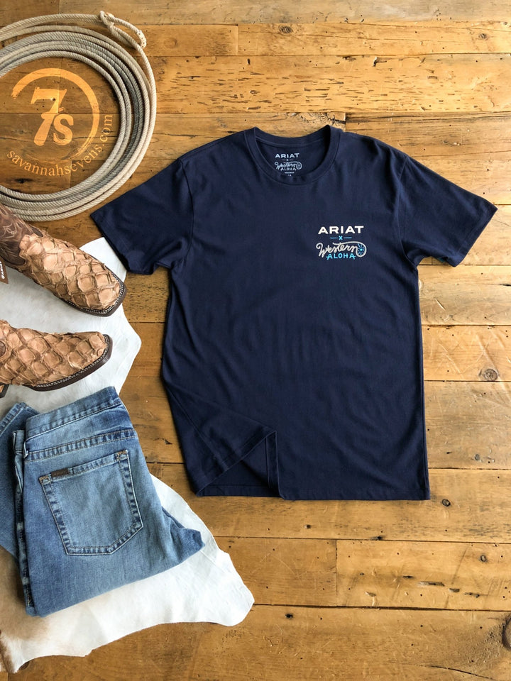 A Pua'a Ariat Men's Graphic Tee by Savannah Sevens western life{&}style in navy blue lies flat on a wooden floor beside brown sandals and folded blue jeans, featuring white printed chest text.