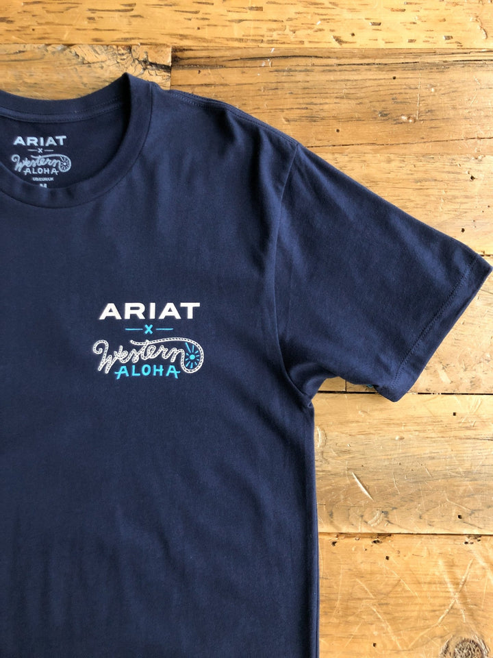 A Pua'a Ariat Men's Graphic Tee by Savannah Sevens western life{&}style, in navy blue with a small white and light blue chest design and a Hawaiian back graphic, is laid flat on a rustic wooden surface.