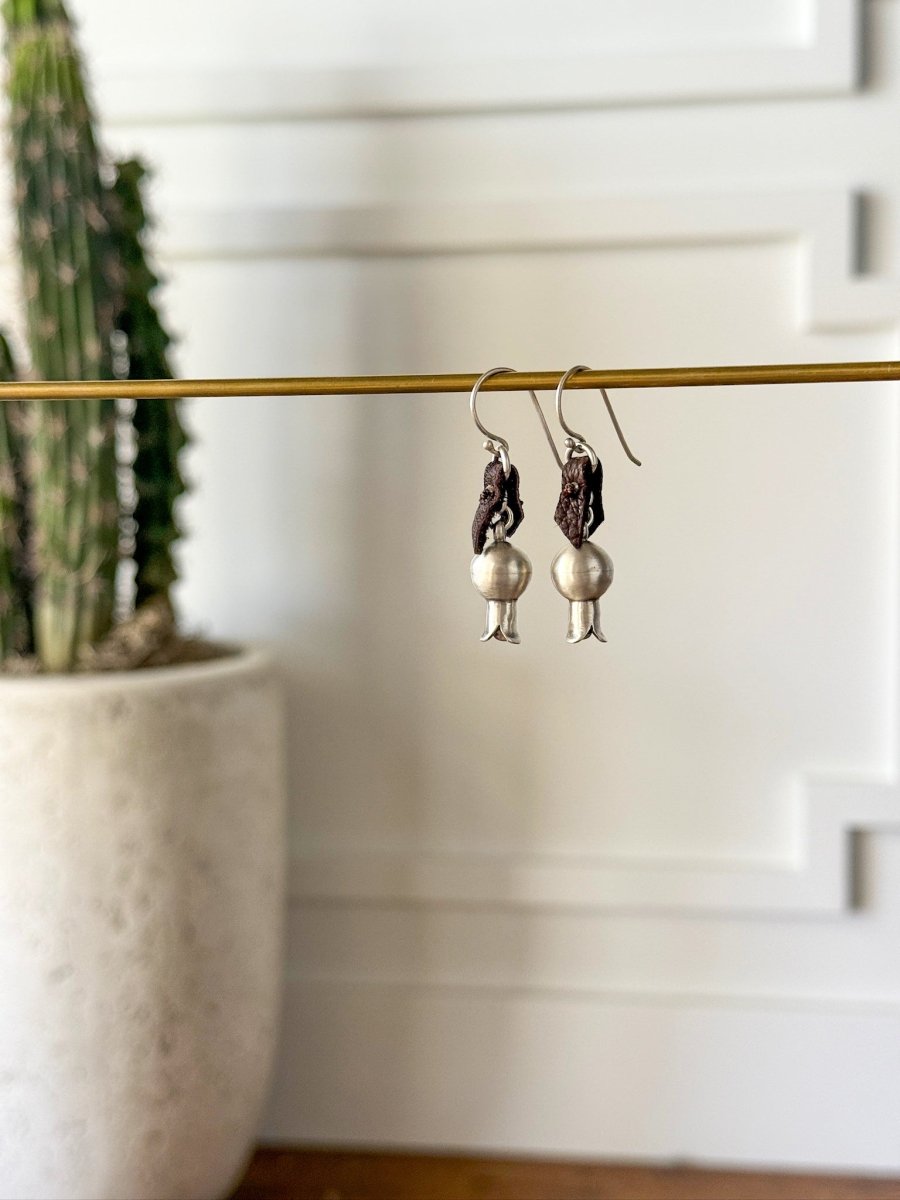 The Puebla Earrings by Savannah Sevens western life{&}style, crafted in sterling silver and dark brown, hang from a gold rod with a blurred potted cactus and white paneled walls in the background.