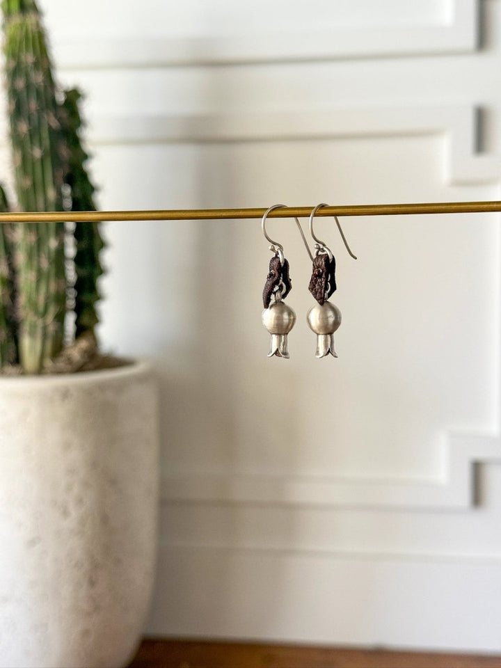 The Puebla Earrings by Savannah Sevens western life{&}style, crafted in sterling silver and dark brown, hang from a gold rod with a blurred potted cactus and white paneled walls in the background.