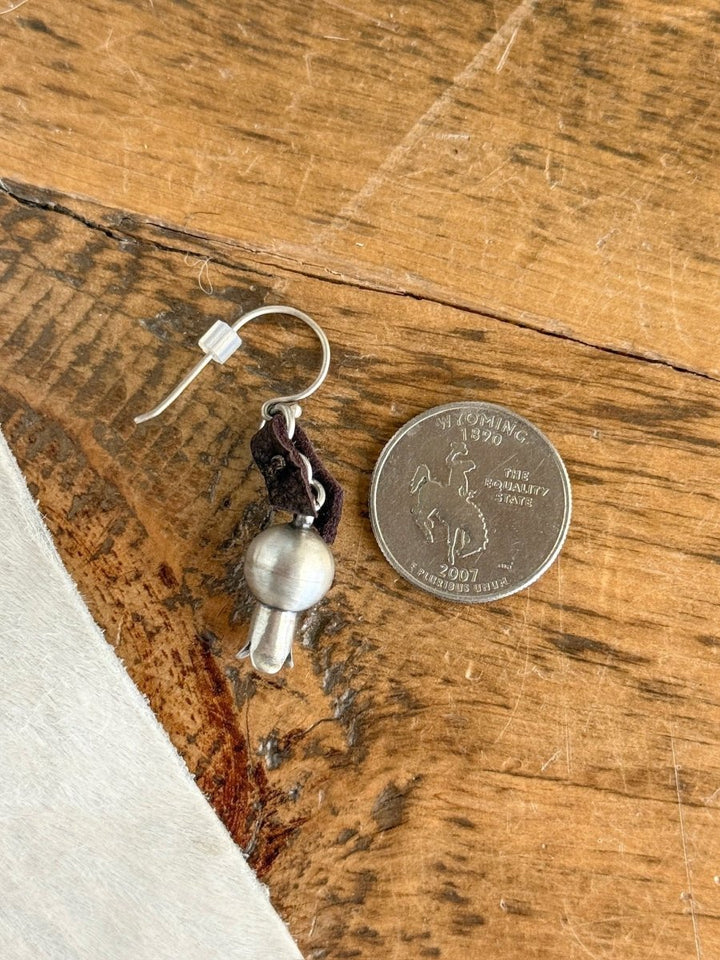 The Puebla Earrings by Savannah Sevens western life{&}style—a sterling silver earring with a round bell and dark loop—rests beside a Wyoming state quarter, showcasing its petite size on a wooden surface.