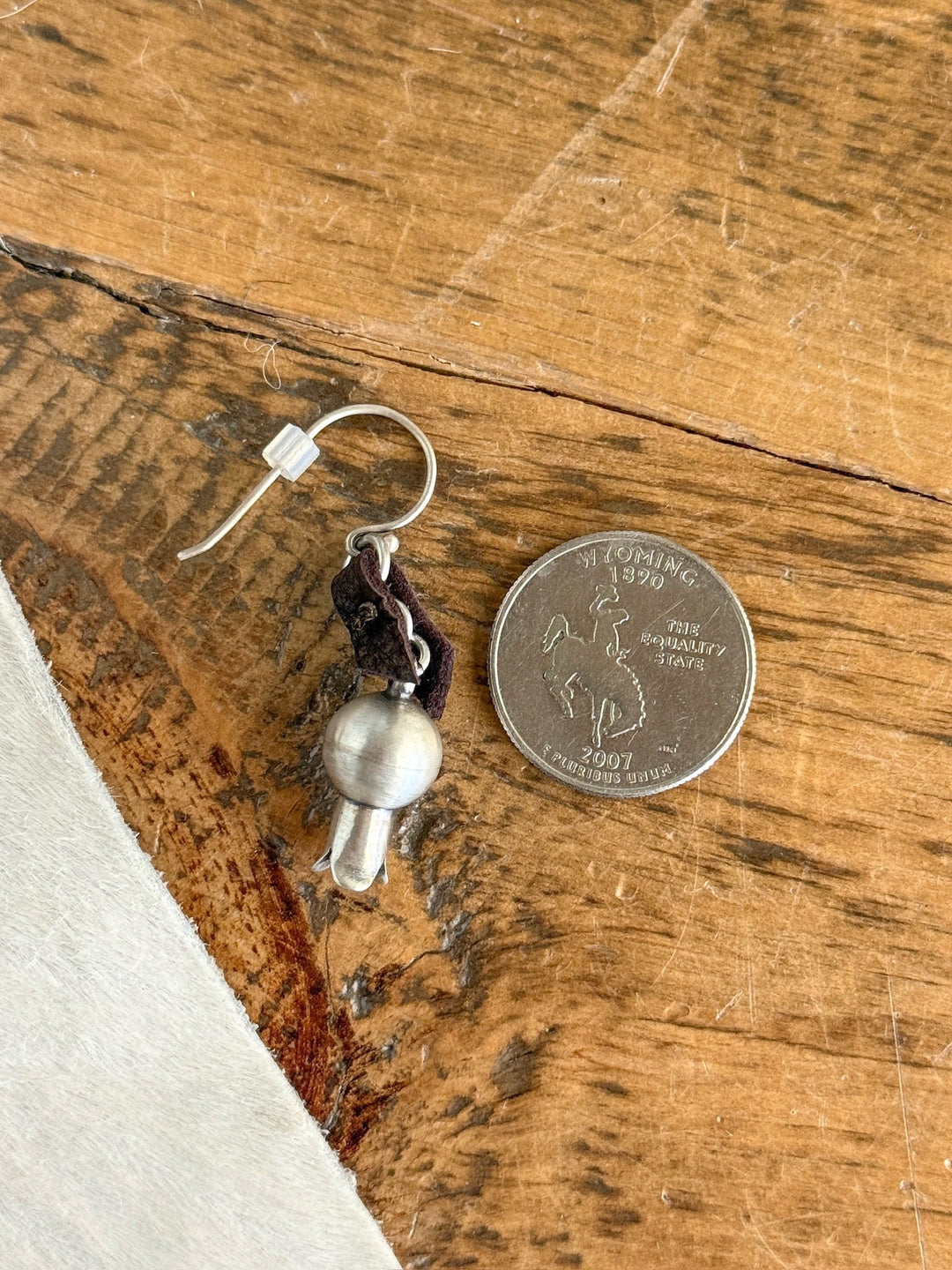 The Puebla Earrings by Savannah Sevens western life{&}style—a sterling silver earring with a round bell and dark loop—rests beside a Wyoming state quarter, showcasing its petite size on a wooden surface.