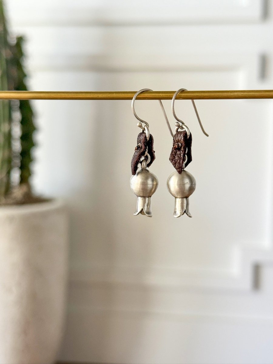 The Puebla Earrings by Savannah Sevens western life{&}style, featuring sterling silver squash blossoms with bell-shaped ends and dark brown accents, hang from a gold rod with a blurred potted plant and white wall in the background.