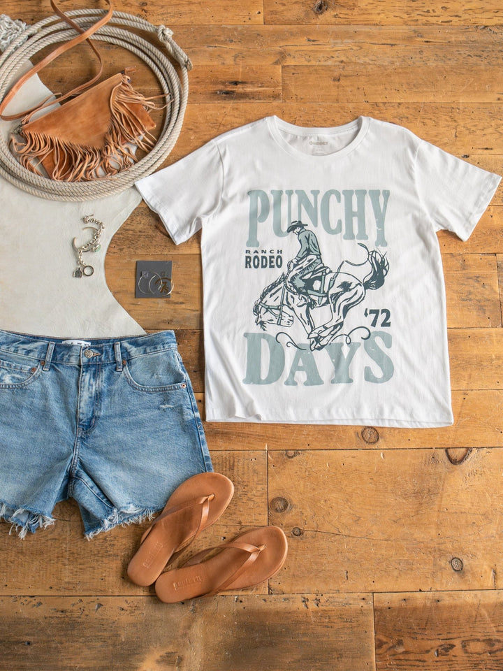 Flat lay of a casual outfit with the Savannah Sevens western life{&}style Punchy Days Graphic Tee, denim shorts, tan sandals, fringed crossbody bag, silver necklace, and coiled rope on a wooden floor.
