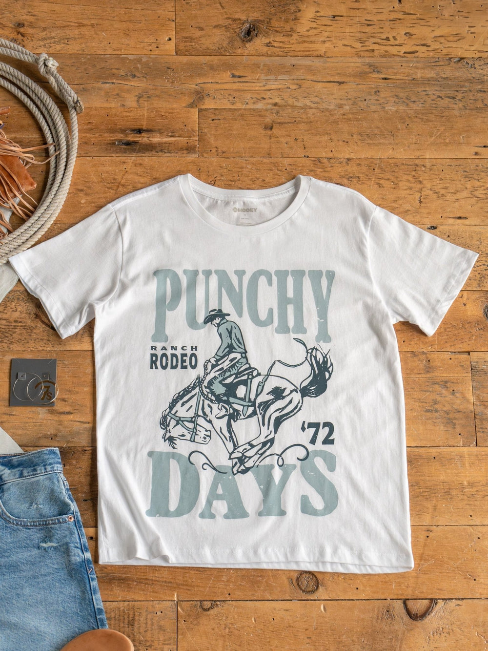 The Punchy Days Graphic Tee by Savannah Sevens western life{&}style, made from 100% cotton with a unisex fit, features a rodeo rider and “PUNCHY DAYS"  graphic. Shown on a wooden floor with denim jeans and a rope nearby.