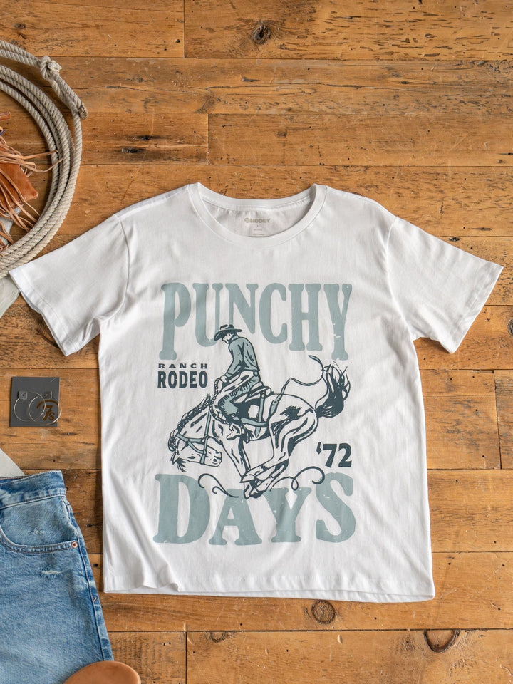 The Punchy Days Graphic Tee by Savannah Sevens western life{&}style, made from 100% cotton with a unisex fit, features a rodeo rider and “PUNCHY DAYS"  graphic. Shown on a wooden floor with denim jeans and a rope nearby.