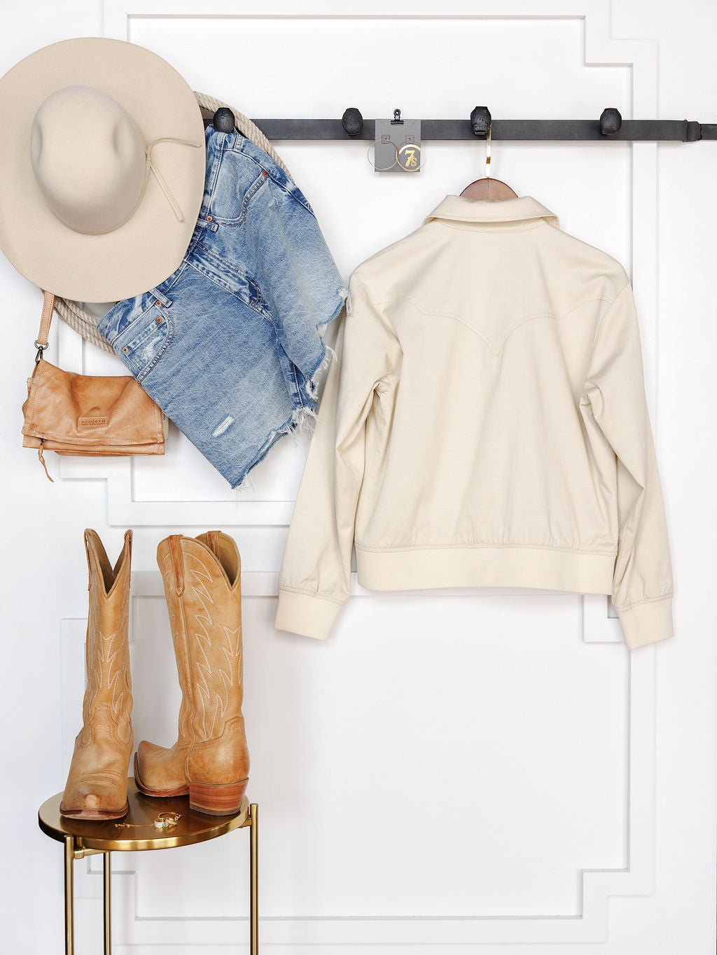 The Questa Boot Stitch Jacket by Savannah Sevens western life{&}style, a tan cowboy hat, denim shorts, and a small tan purse are hung above a gold stool with embroidered tan cowboy boots, set against a white paneled wall.
