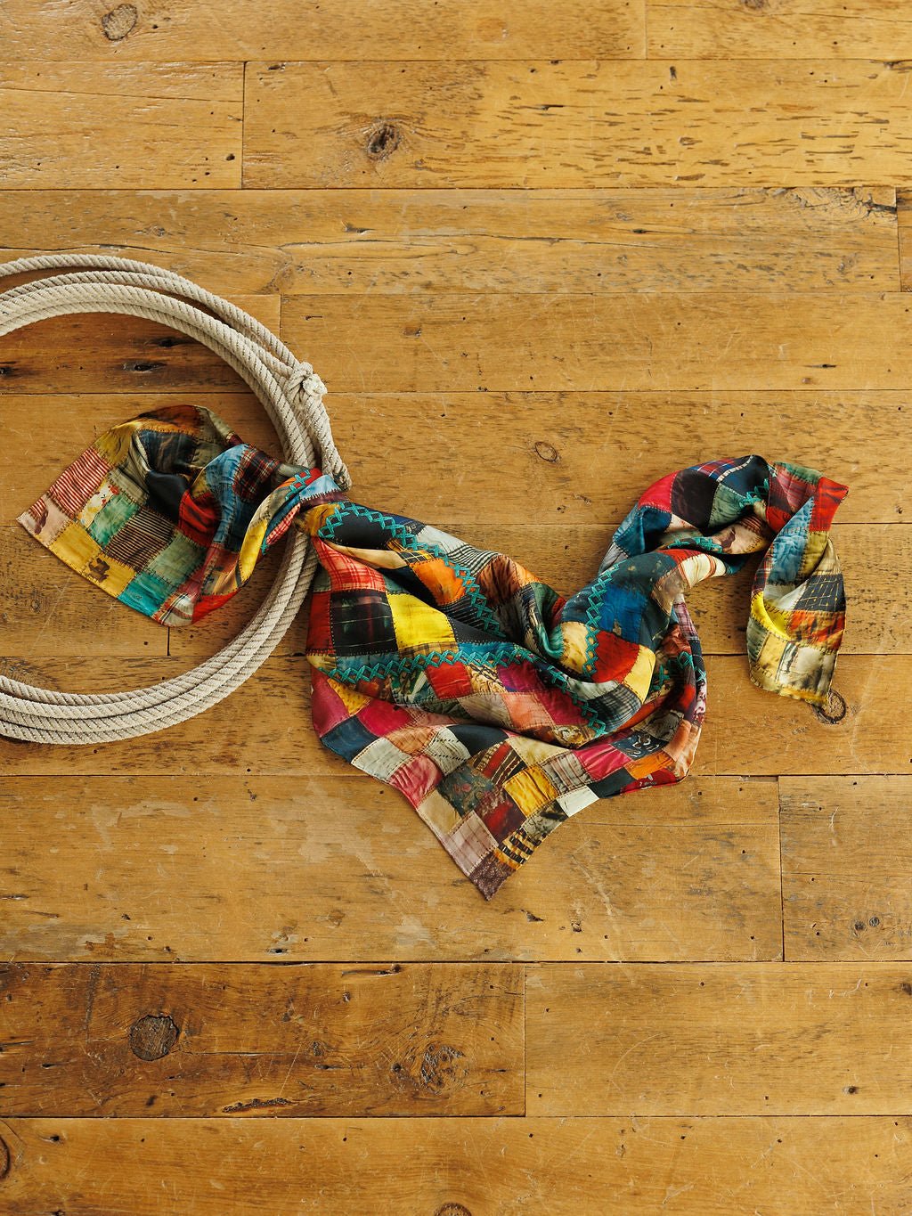A multi-colored Quilt Silk Scarf from Savannah Sevens western life&style laid on a wooden floor next to a coiled rope.